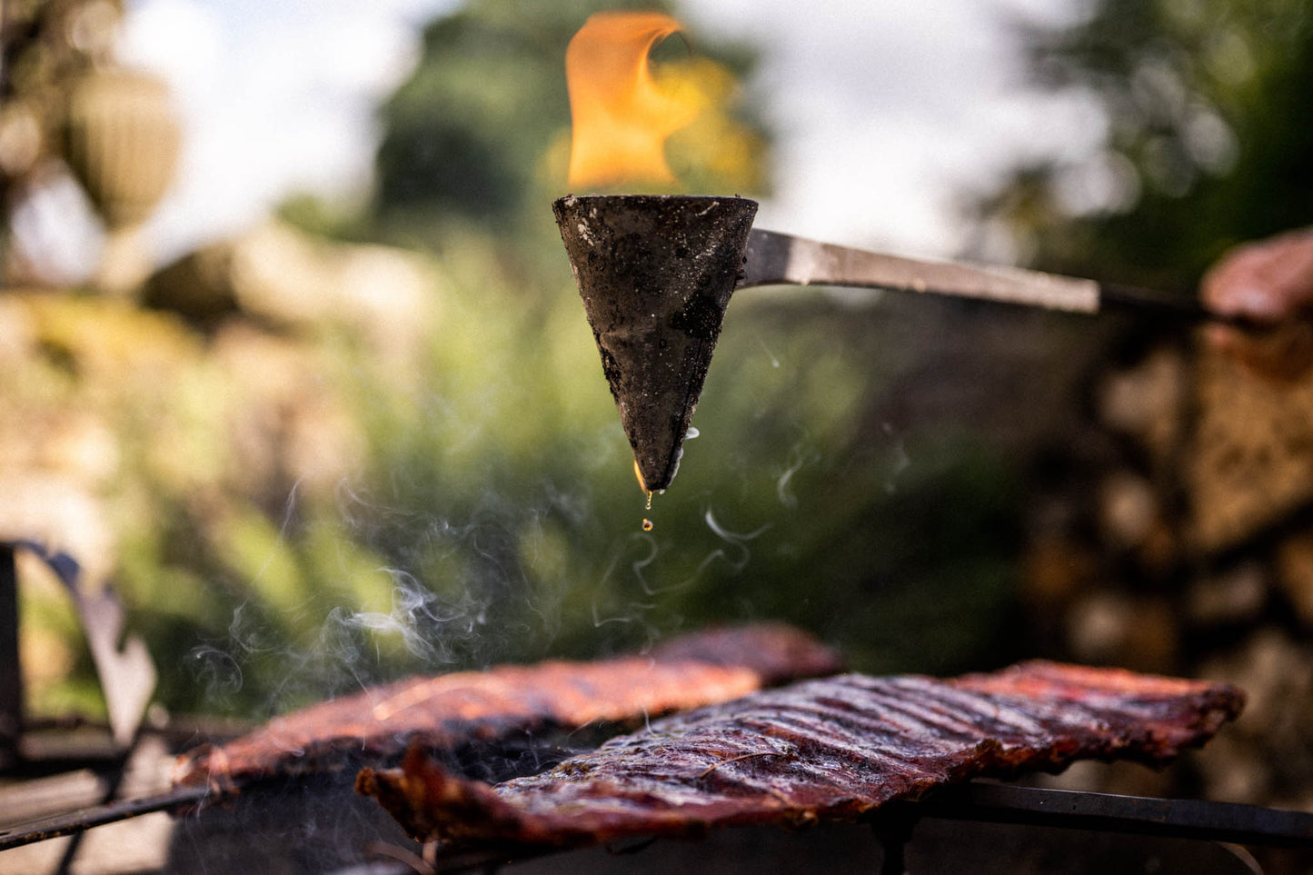 Flaming flambadou suspended above smoked ribs, dripping hot rendered fat onto meat over an outdoor grill