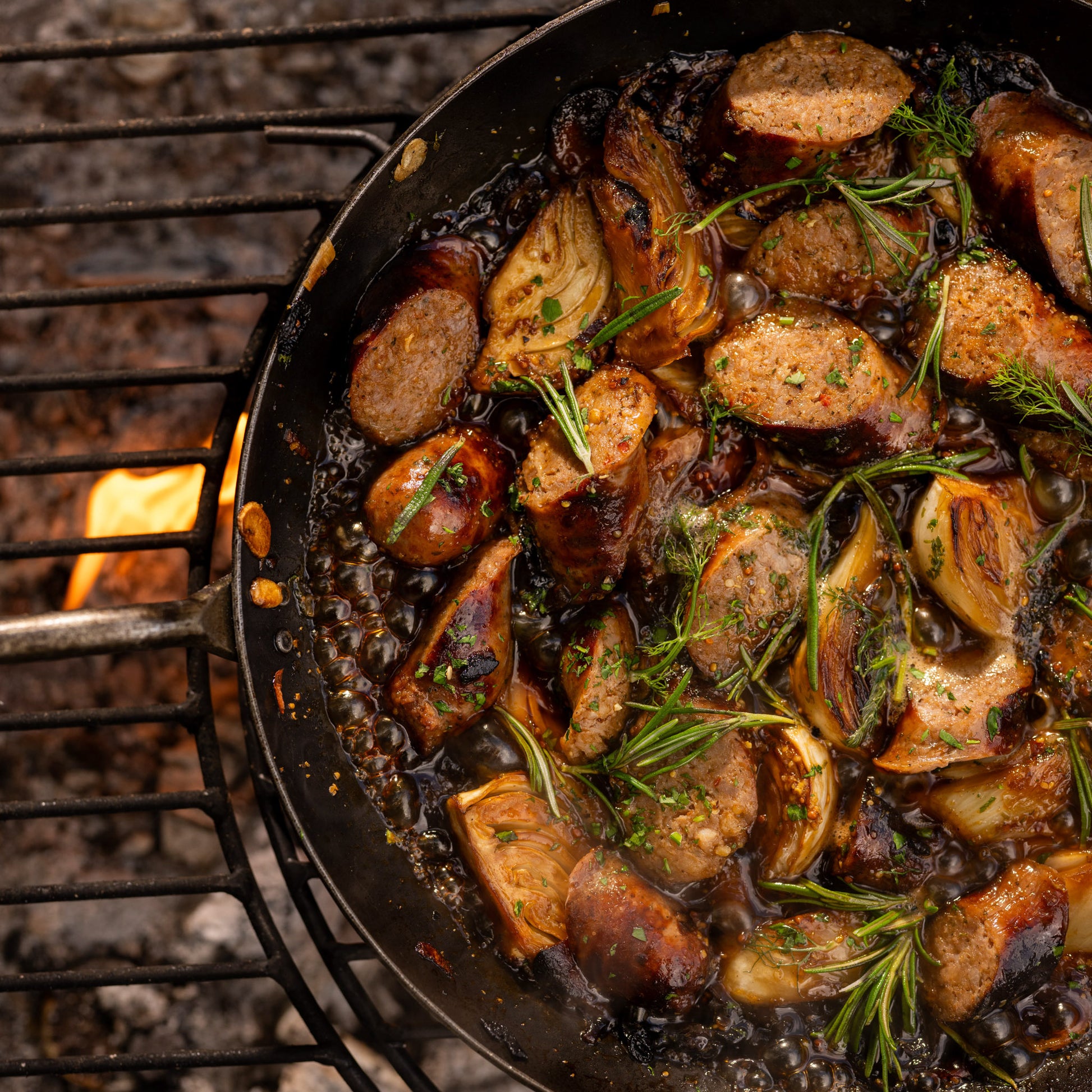 Close-up of a BBQ frying pan with sausages, caramelised onions and rosemary cooking over flames on a Kadai firebowl barbecue