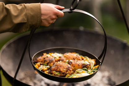Tripod skillet hanging over a Kadai firebowl barbecue with roasted chicken and vegetables being cooked outdoors over hot coals