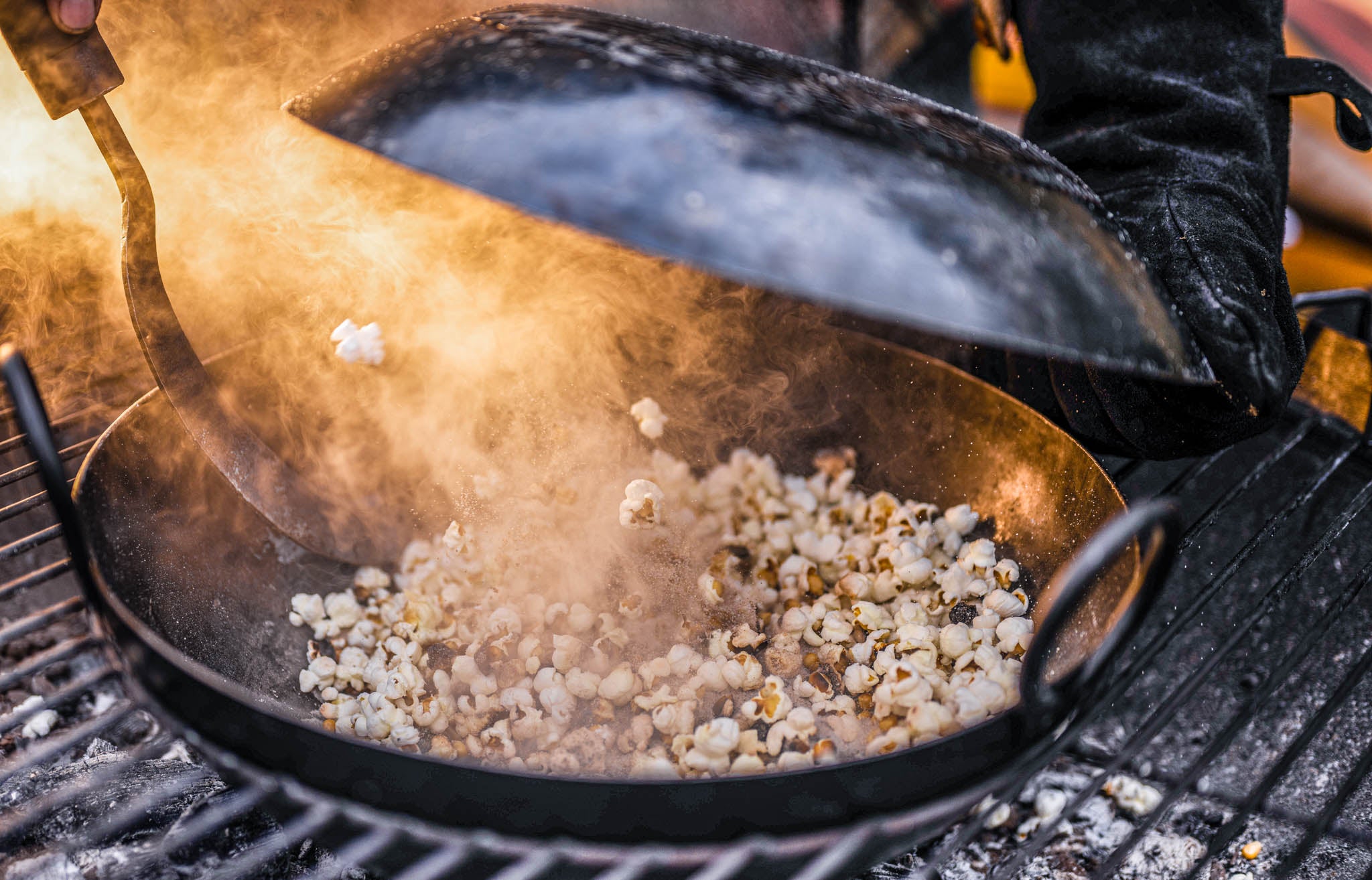 Outdoor popcorn maker in action with popcorn cooking over hot coals in a rustic fire pit setup