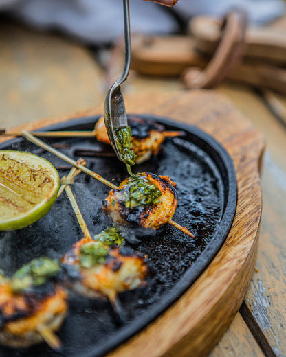 Hands adding green sauce on grilled shrimp skewers on a hot sizzler plate with lime.