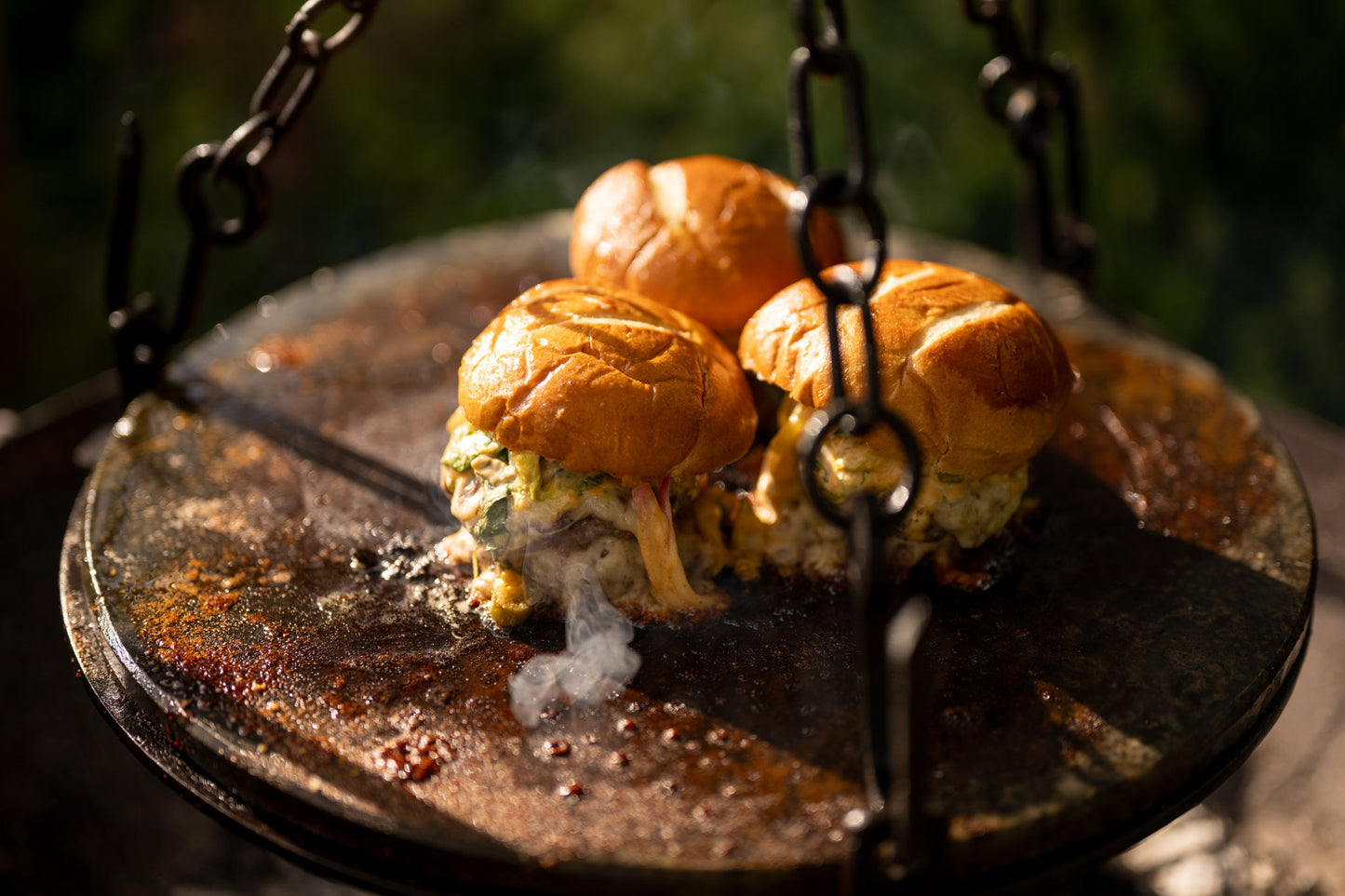 Stone griddle plate suspended over a Kadai firebowl barbecue with sizzling cheeseburgers cooking above hot coals outdoors