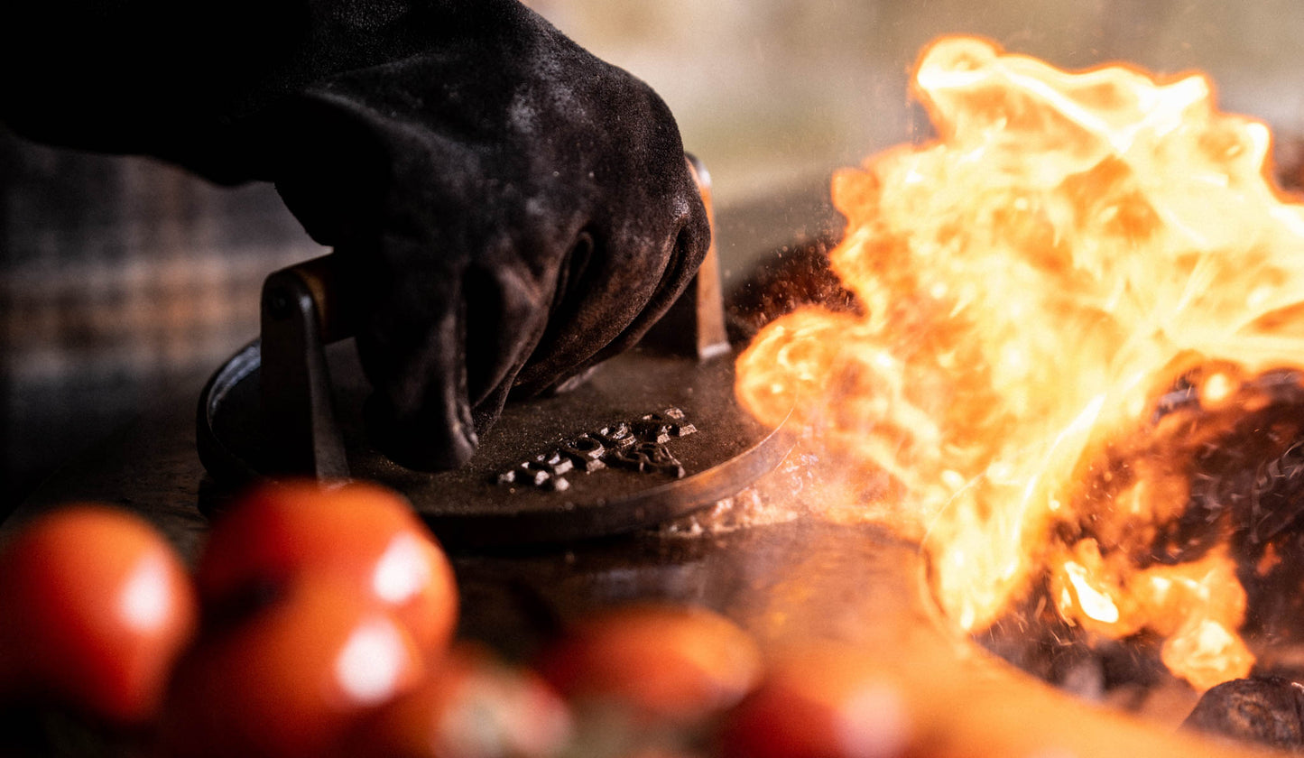Hand with black glove using a burger press on a grill with flames and tomatoes in the foreground.