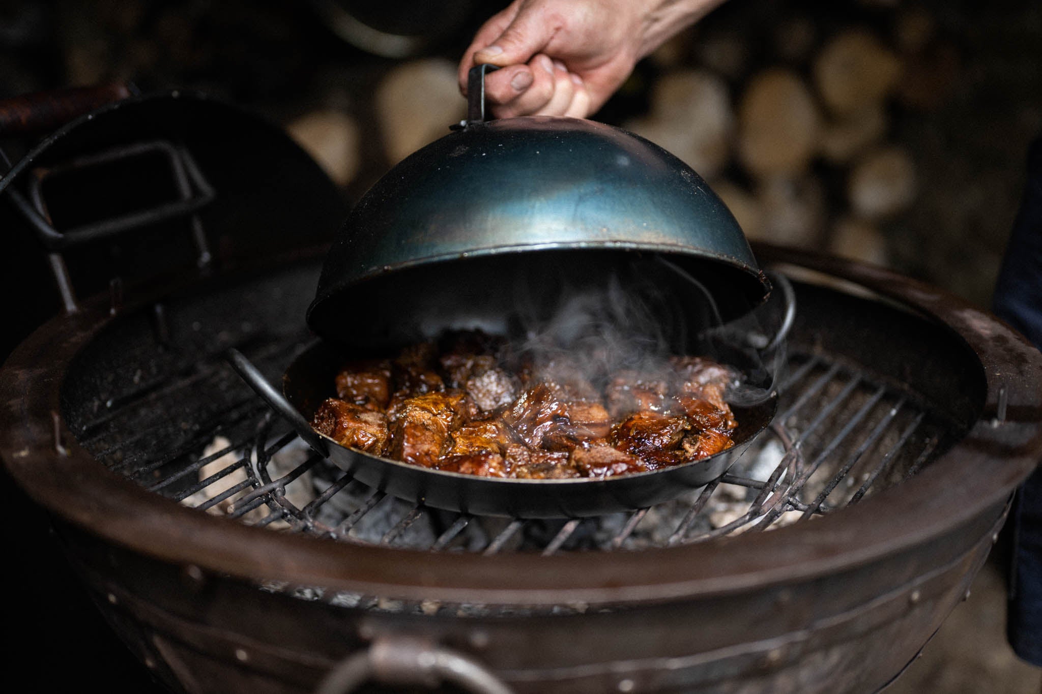 Hand lifting a metal barbecue cloche from a smoking grill pan filled with richly glazed meat on an outdoor fire bowl