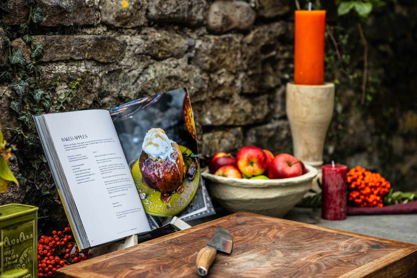 Wooden cutting board with a knife, open book, and fruit bowl against a stone wall.