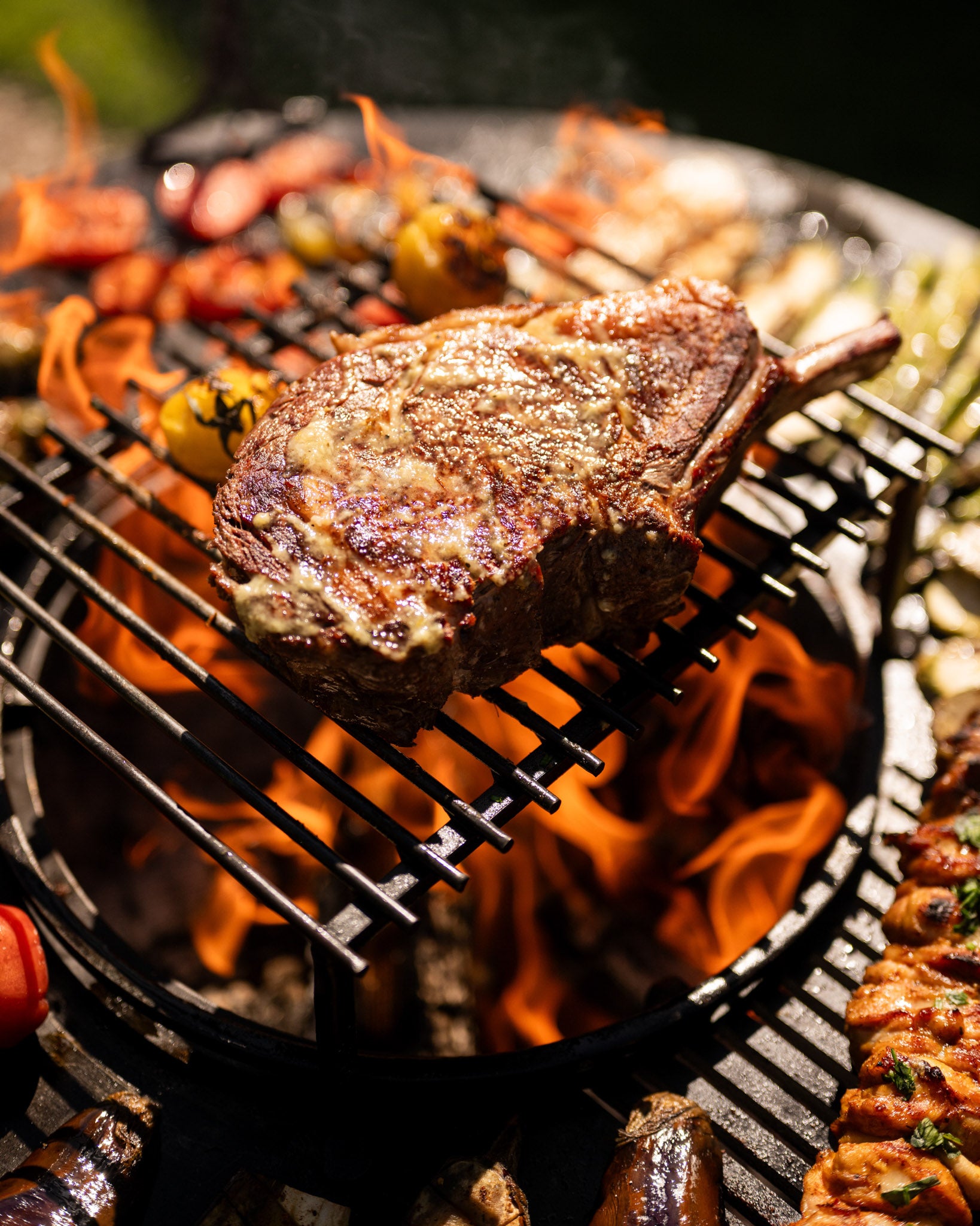 Juicy steak sizzling over open flames on a BBQ cooking grill with grilled vegetables and skewers in the background.