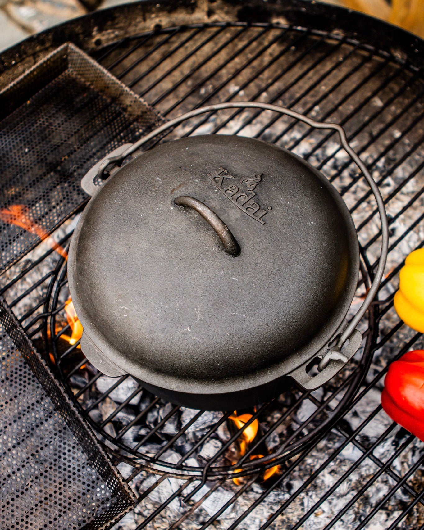 A black cast iron Dutch oven with the brand name 'Kadoi' embossed on top, sitting on a metal grill over coals and flames.