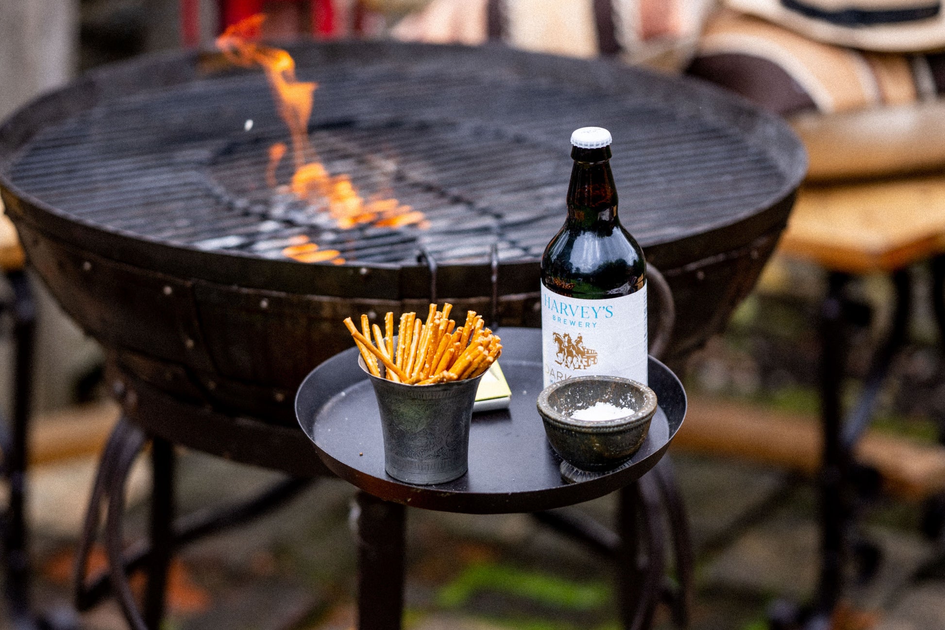 Round black Kadai side shelf attached to an outdoor fire bowl, styled with snacks, seasoning, and a bottled drink beside the grill