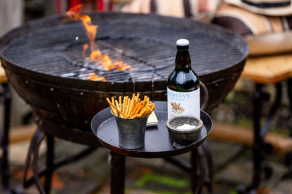 Round black Kadai side shelf attached to an outdoor fire bowl, styled with snacks, seasoning, and a bottled drink beside the grill