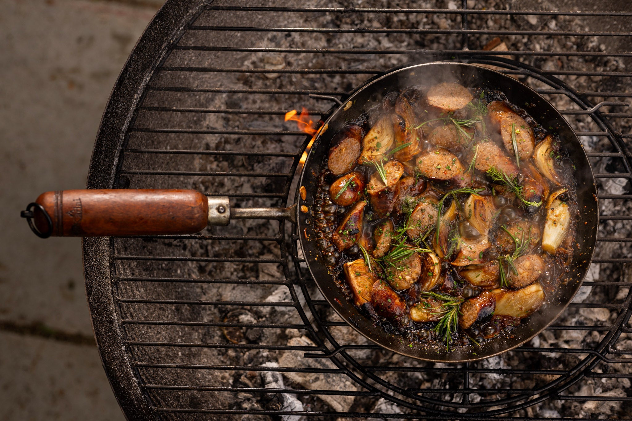BBQ frying pan with sausages, caramelised onions and rosemary cooking over hot coals on a Kadai barbecue firebowl