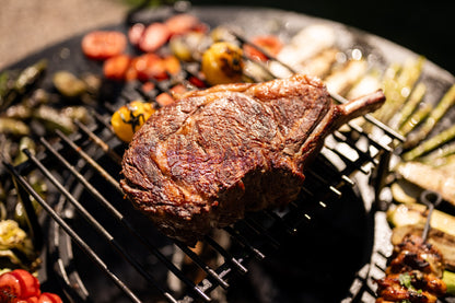 Large grilled steak cooking on a BBQ grill surrounded by roasted vegetables and skewers over open flames.