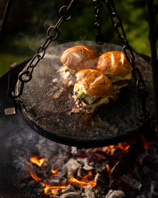 Stone griddle plate suspended over open flames on a Kadai firebowl barbecue with burgers cooking above hot coals outdoors