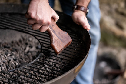 Close-up of a handcrafted wooden BBQ scraper maintaining a charcoal grill grate after cooking outdoors