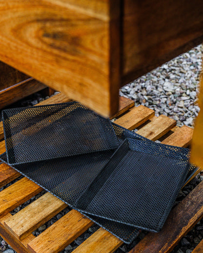 Perforated black BBQ grill trays displayed on a wooden pallet outdoors, designed for grilling vegetables, seafood, and smaller foods over open flames.