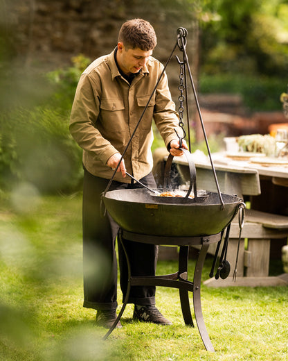 Tripod skillet hanging over a Kadai firebowl barbecue with outdoor cooking taking place in a garden setting