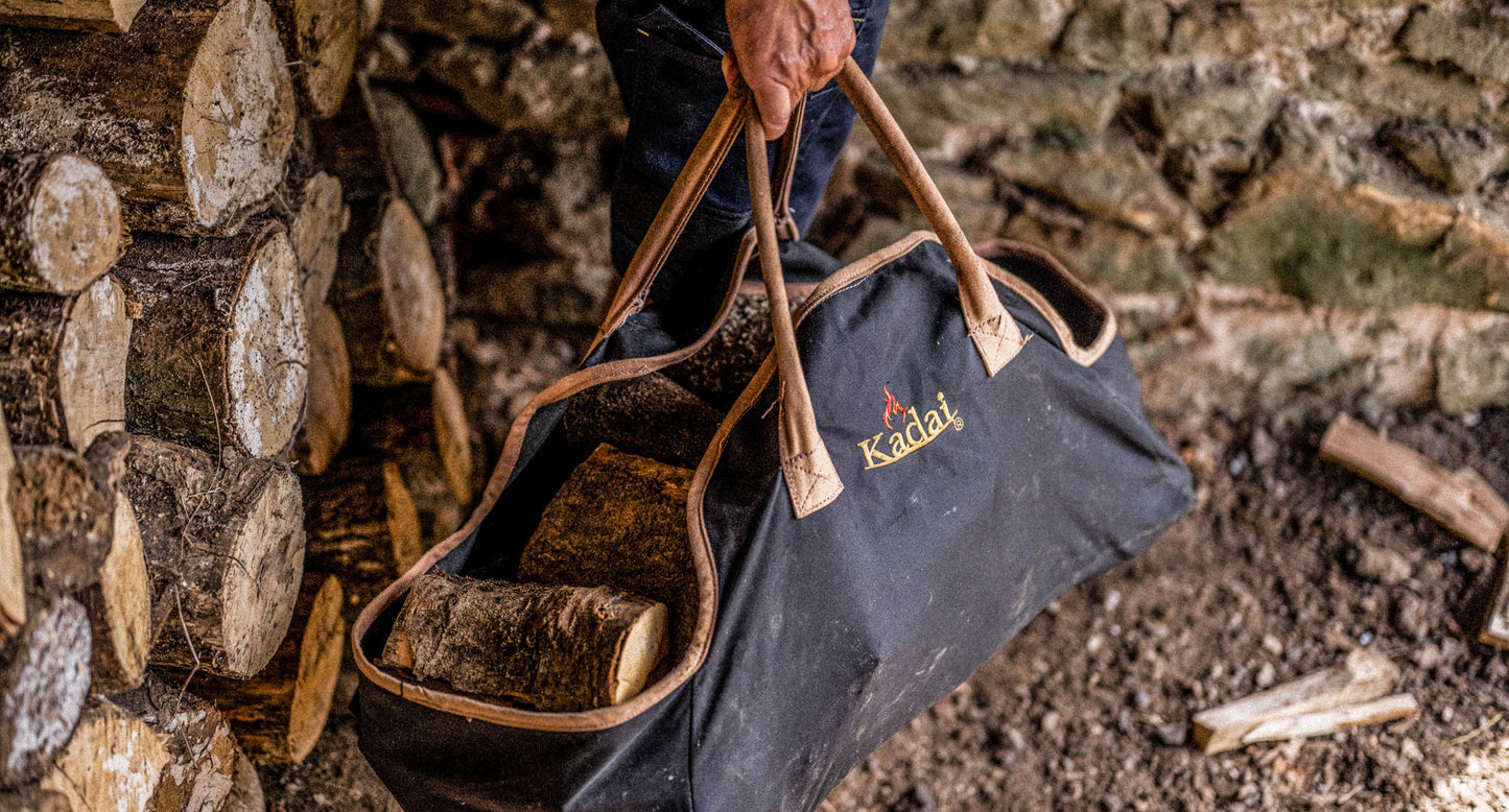 Person carrying firewood logs in a black canvas log carrier bag beside a stacked wood pile.