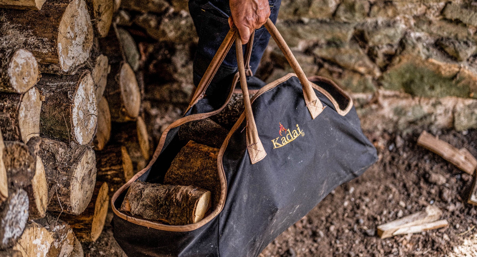 Person carrying firewood logs in a black canvas log carrier bag beside a stacked wood pile.