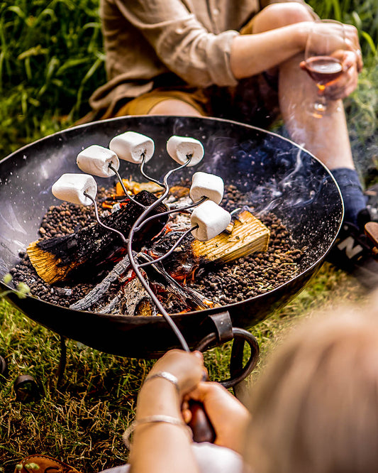 Marshmallows roasting on a long metal fork above a Kadai fire bowl with glowing embers and logs outdoors