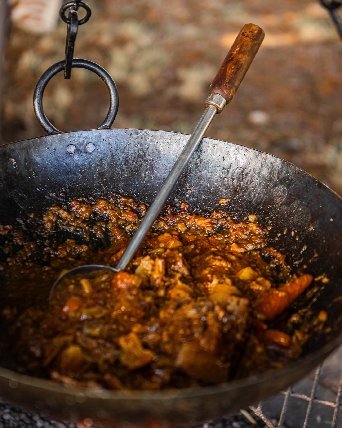Long-handled BBQ ladle resting inside a Kadai fire bowl filled with slow-cooked stew and vegetables