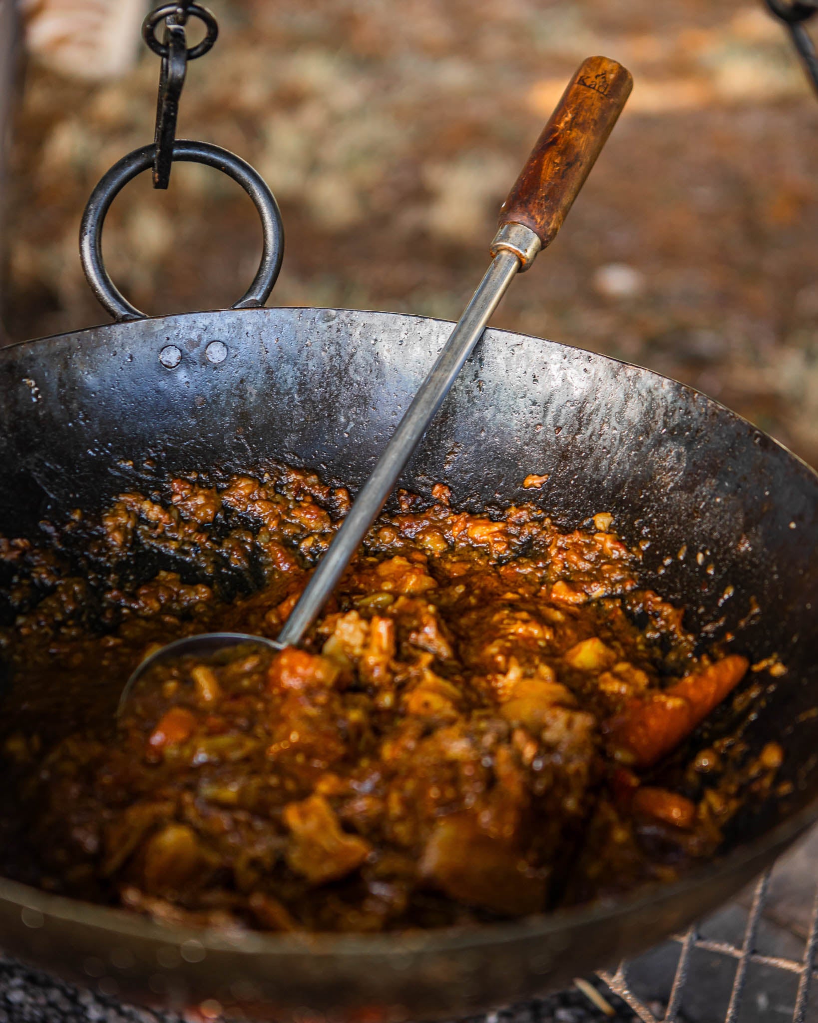Long-handled BBQ ladle resting inside a Kadai fire bowl filled with slow-cooked stew and vegetables