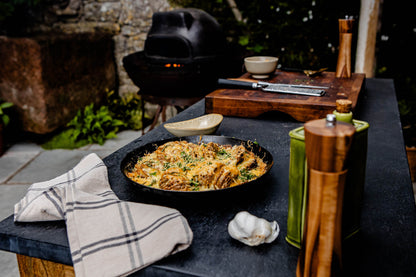 Baked oven dish in a skillet topped with melted cheese and herbs, placed on an outdoor countertop with garlic, oil bottle, and cooking tools nearby