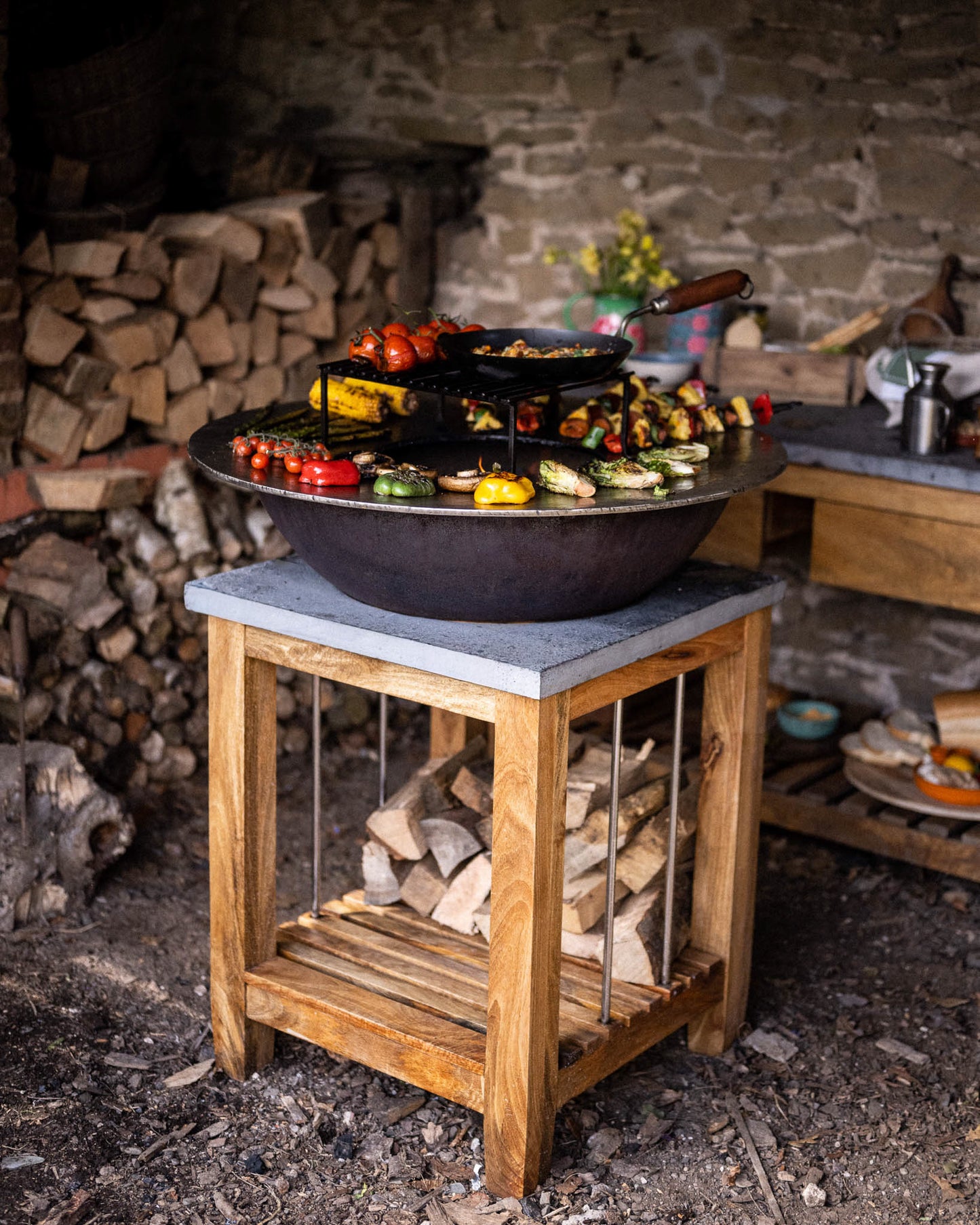 Outdoor BBQ plancha station with a circular flat-top grill on a wooden stand, surrounded by grilled vegetables, skewers, and cookware in a rustic cooking area.