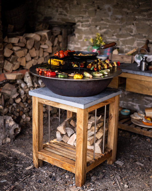 Outdoor BBQ plancha station with a circular flat-top grill on a wooden stand, surrounded by grilled vegetables, skewers, and cookware in a rustic cooking area.