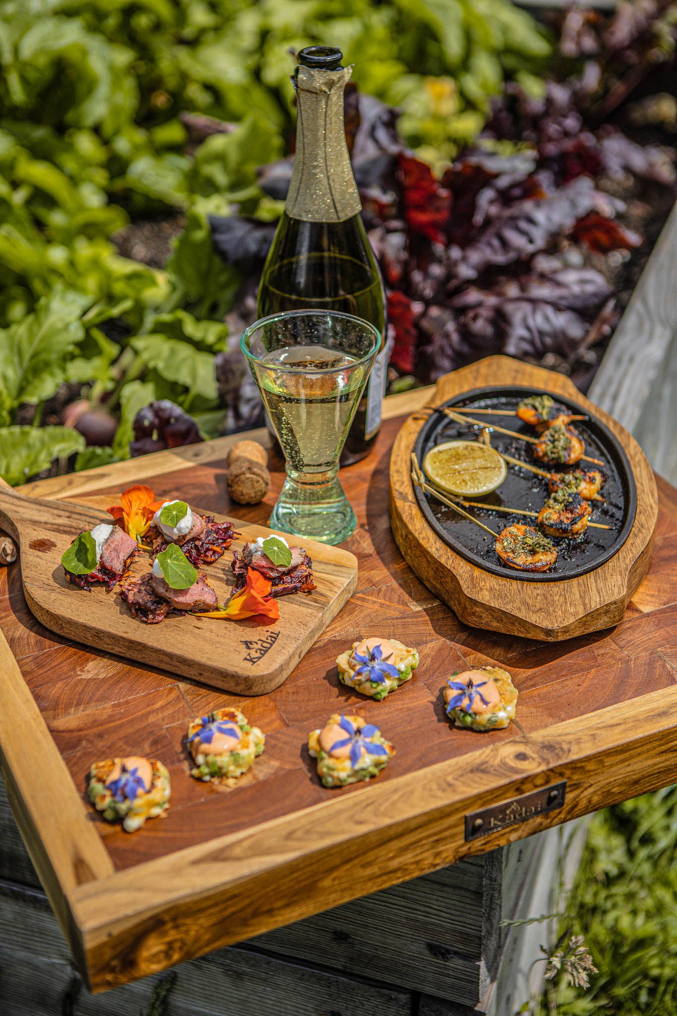 Rustic butcher’s block serving board displaying gourmet appetizers, grilled seafood, and sparkling wine beside a lush garden backdrop.