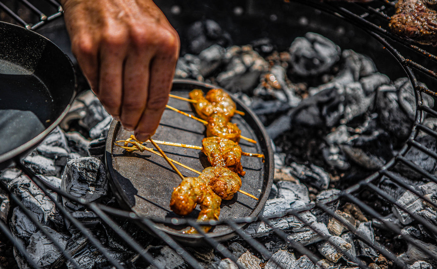 Hands adjusting skewers of marinated meat on a sizzling plate over charcoal.