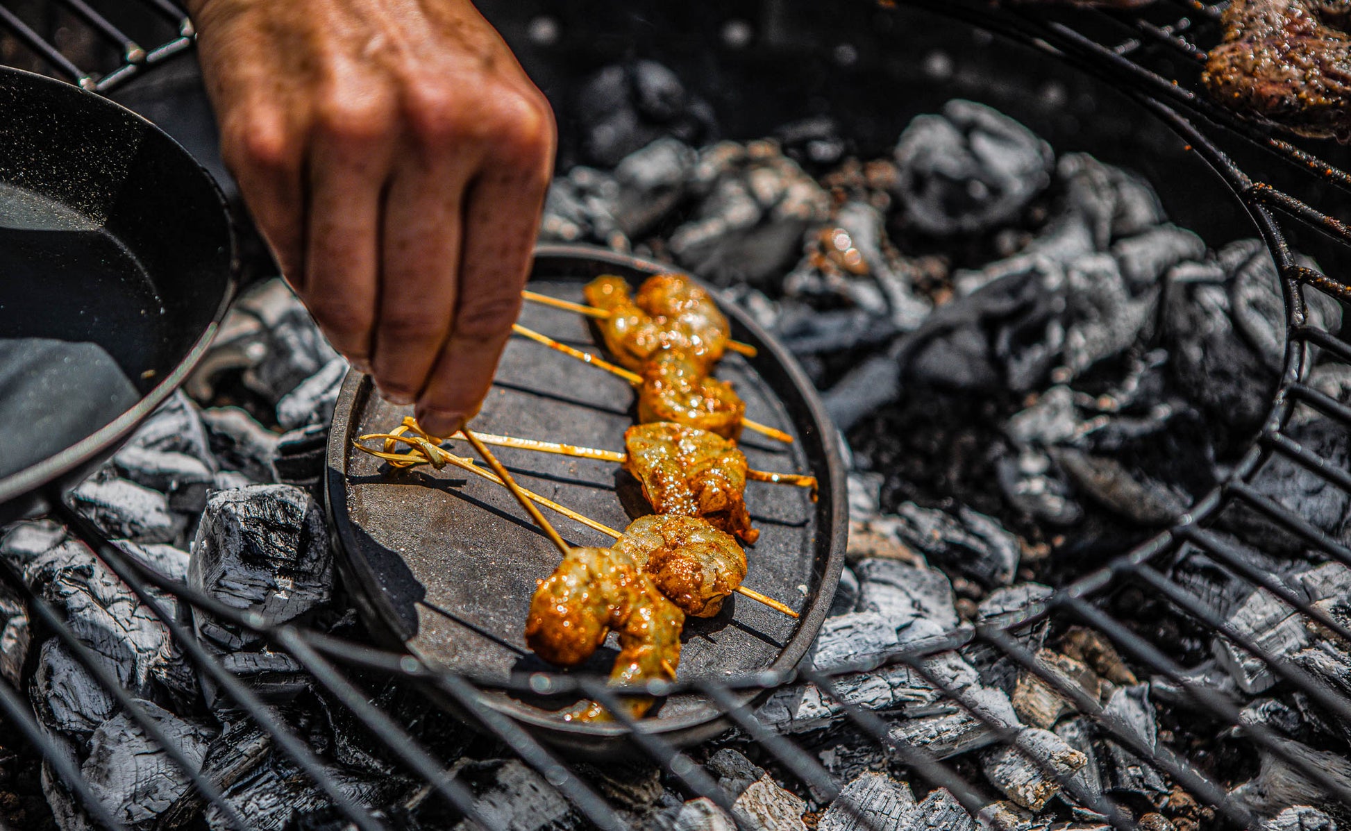 Hands adjusting skewers of marinated meat on a sizzling plate over charcoal.