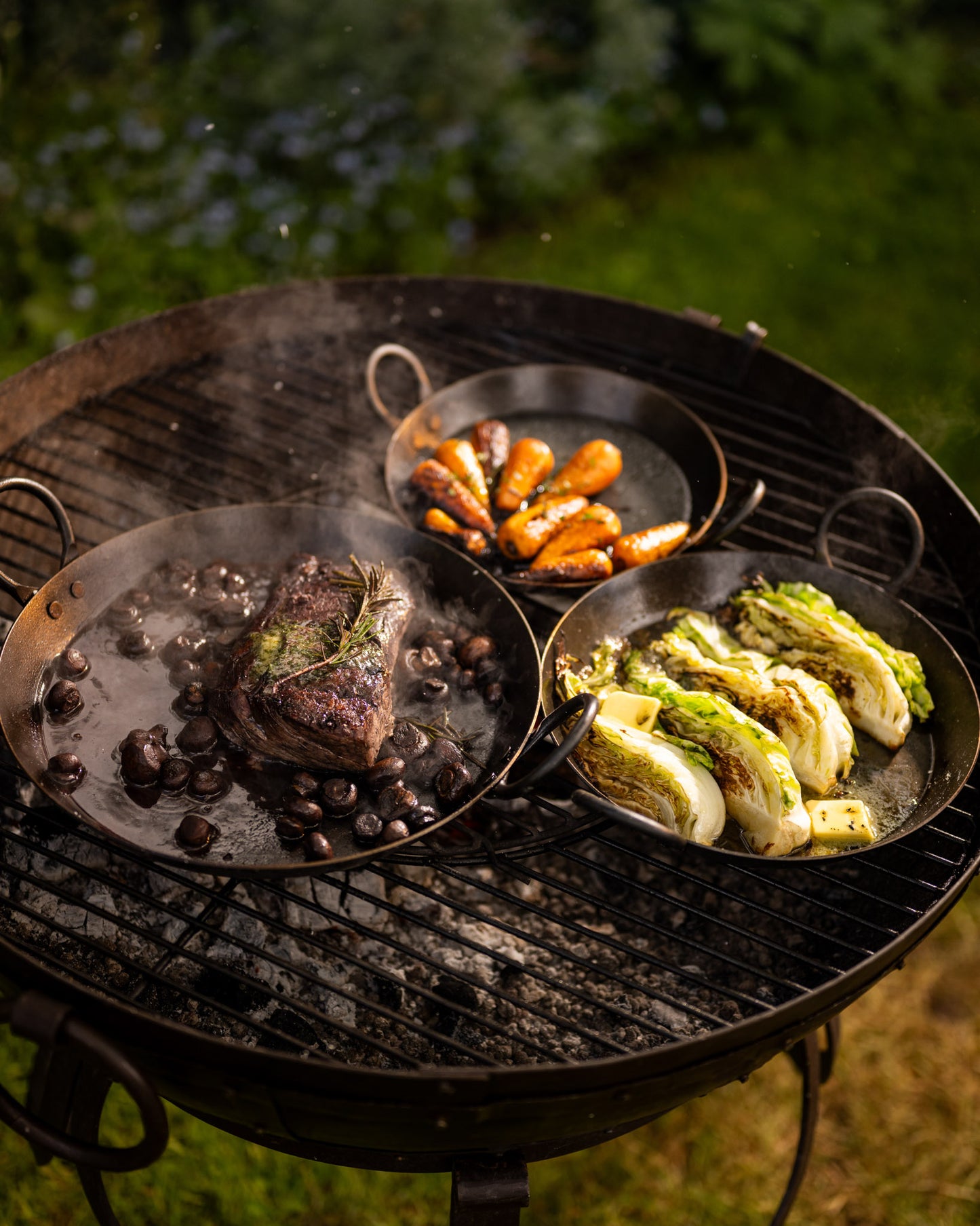 BBQ skillets cooking steak, mushrooms, roasted carrots, and charred cabbage over hot coals on an outdoor grill.