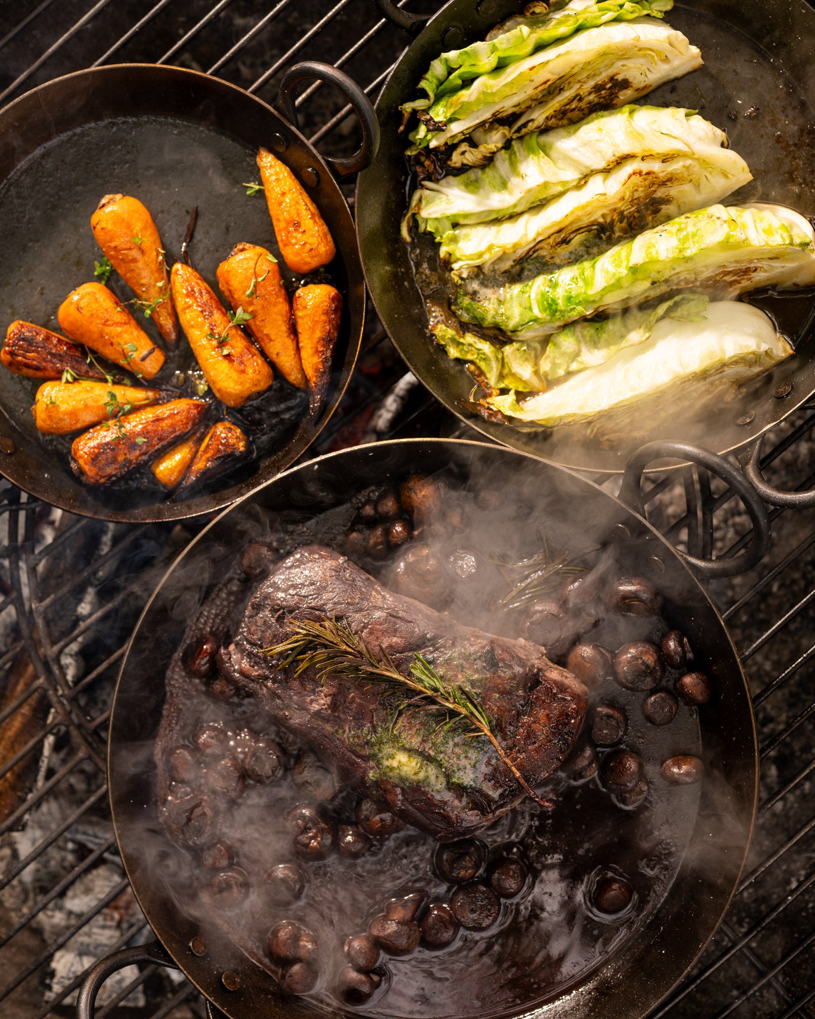 “BBQ skillets filled with grilled steak, roasted carrots, charred cabbage, and mushrooms cooking over hot coals and open flames.