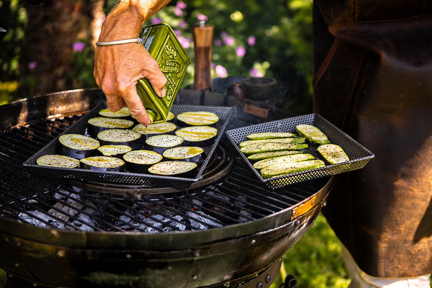 Person seasoning sliced vegetables on perforated BBQ grill trays over a charcoal grill outdoors.