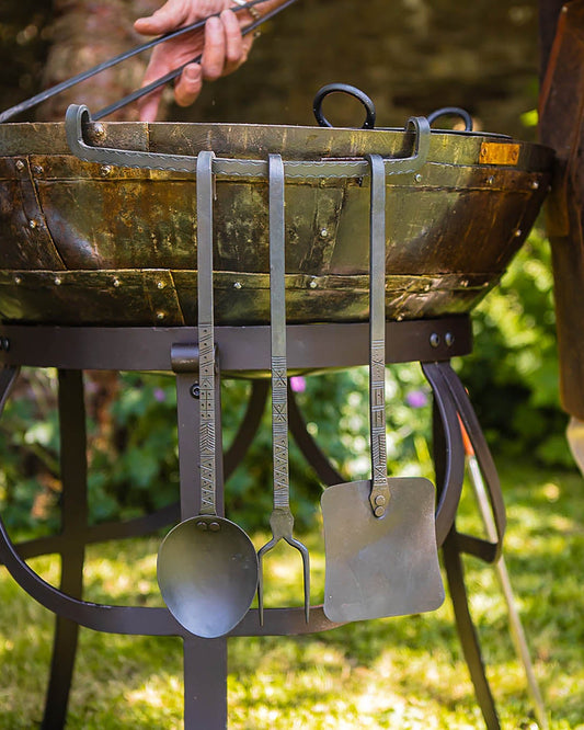 Traditional metal cooking utensils hanging from a rustic outdoor fire bowl while a person tends the fire in a garden setting.
