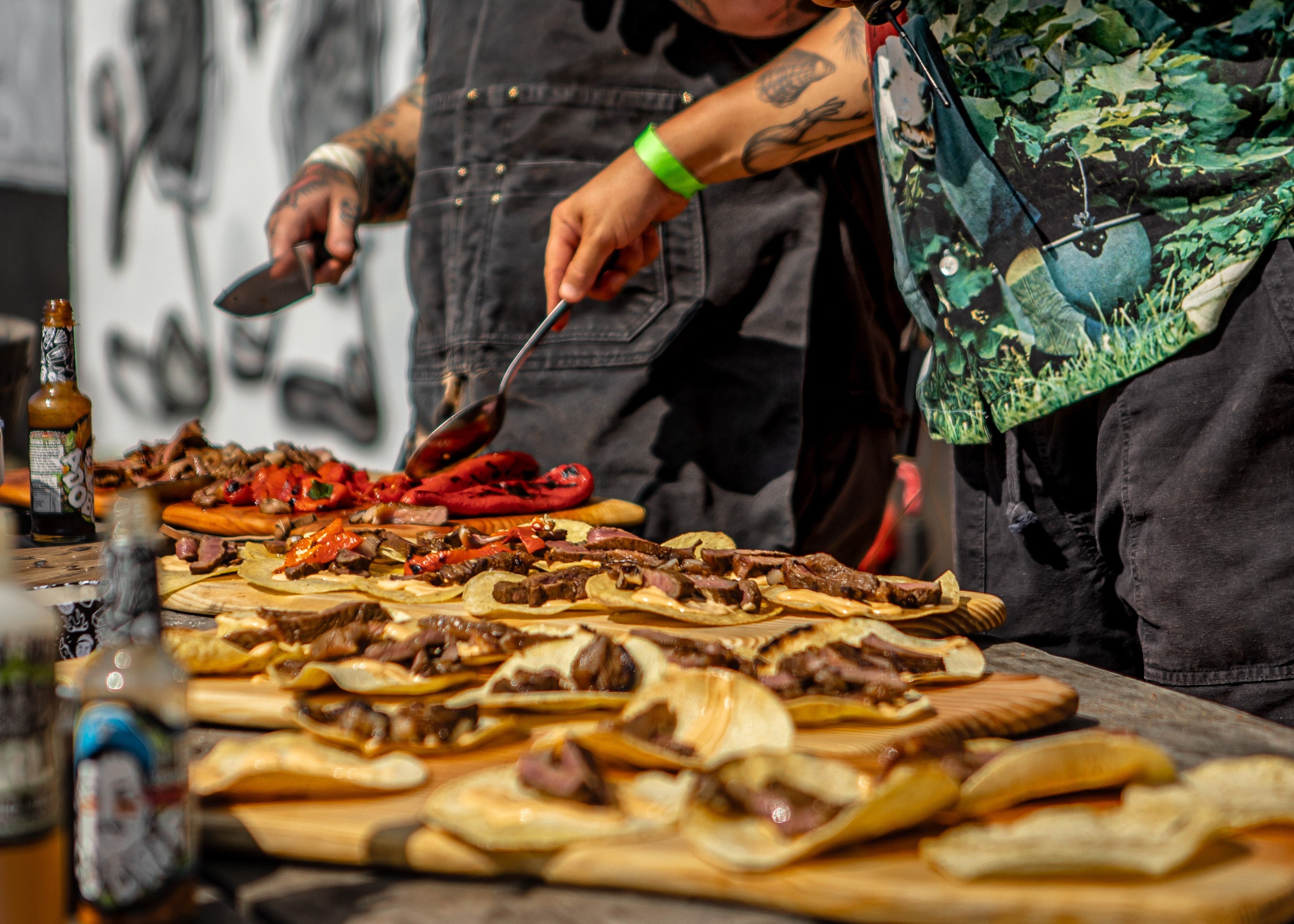 Two men preparing food on a grill outdoors with bottles and ingredients around.