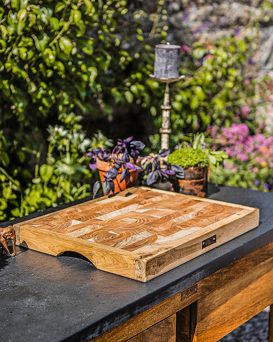 Thick wooden butcher’s block chopping board displayed on an outdoor prep table with garden plants in the background.