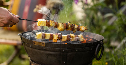 Skewers of pineapple and sausage being lifted from a portable BBQ skillet over open flames