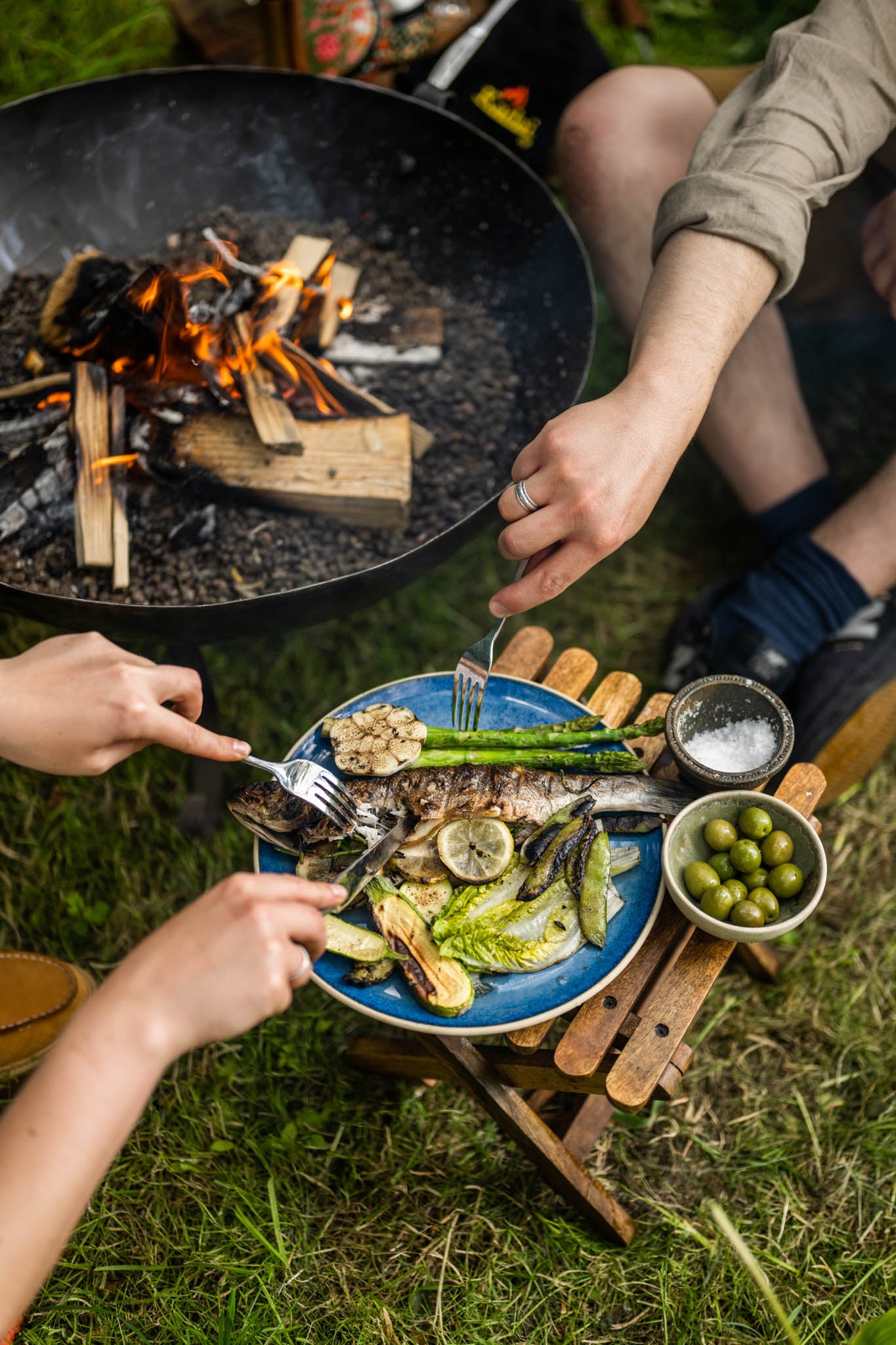 Foldable camping stool used as a table beside a fire pit with grilled fish and vegetables