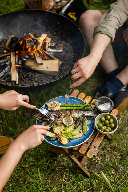 Foldable camping stool used as a table beside a fire pit with grilled fish and vegetables