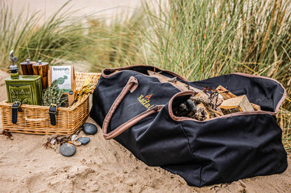 Black canvas log carrier filled with firewood beside a picnic basket on sandy ground surrounded by coastal grasses.