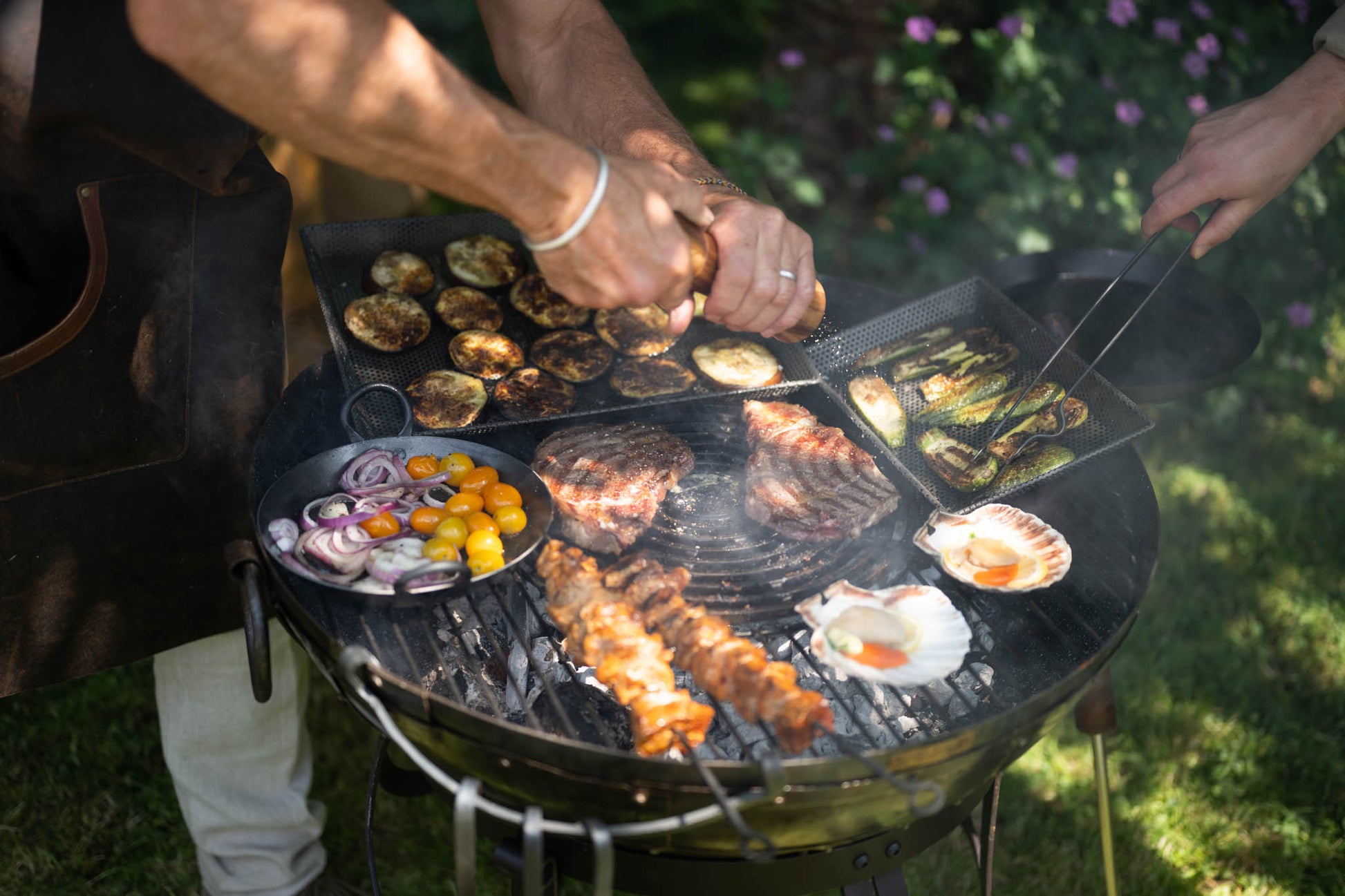 Outdoor barbecue cooking setup with meats, vegetables, and seafood grilling on a stone griddle plate over charcoal