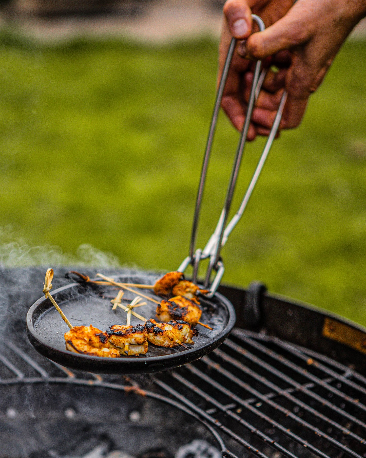 Hand using tongs to lift skewered grilled shrimp from a hot sizzler plate over a charcoal grill.