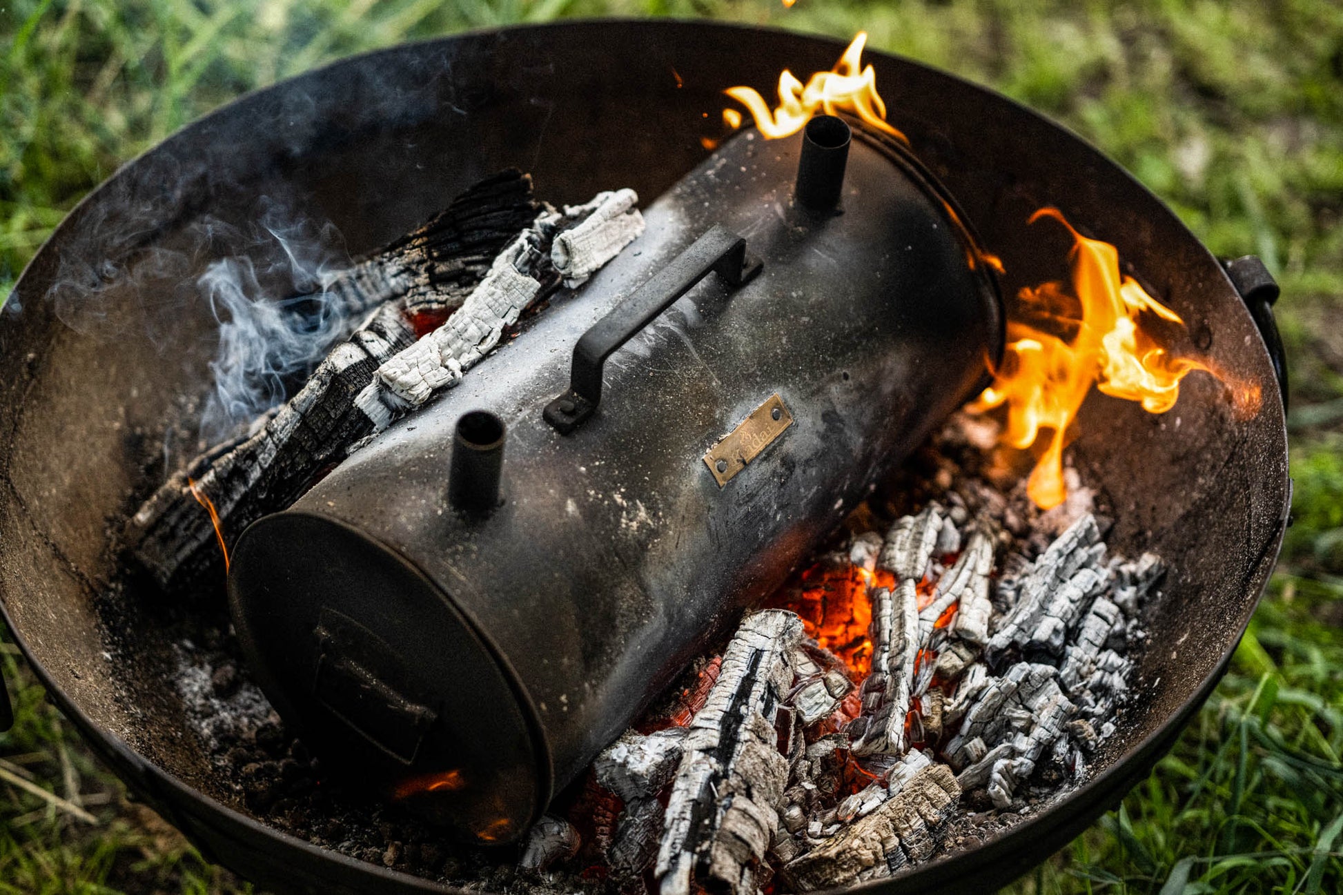 Black metal charcoal maker resting among glowing embers and flames inside a Kadai fire bowl, with smoke rising from charred logs outdoors.