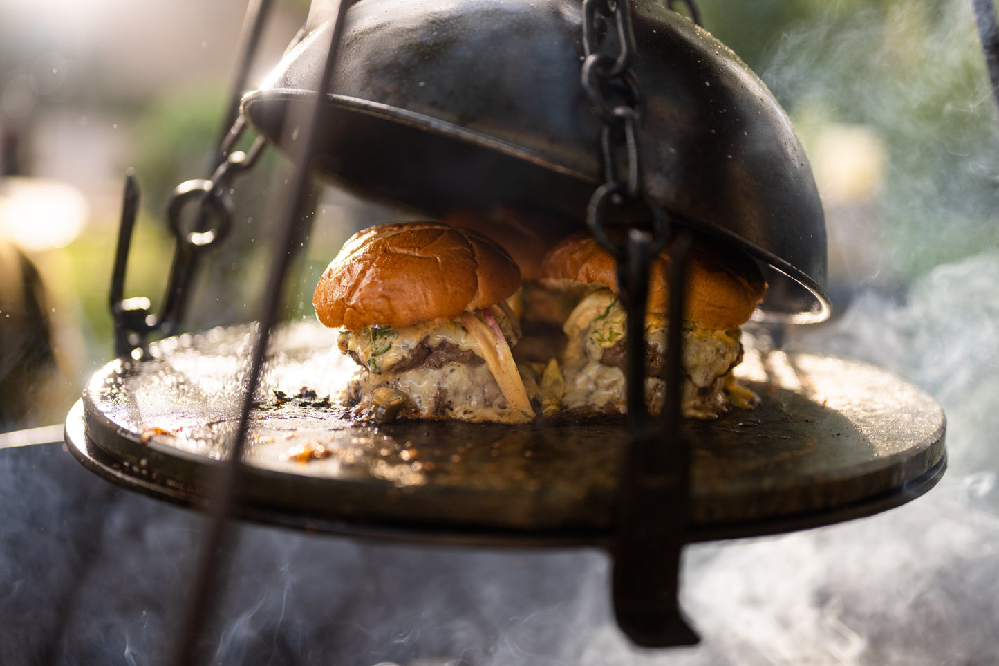 Cheeseburgers steaming beneath a BBQ cloche on a hot stone griddle plate over an outdoor fire cooking setup.