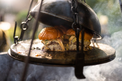 Cheeseburgers steaming beneath a BBQ cloche on a hot stone griddle plate over an outdoor fire cooking setup.