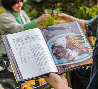 Open Kadai live fire cookbook showing steak recipe with grilled meat and vegetables beside outdoor barbecue