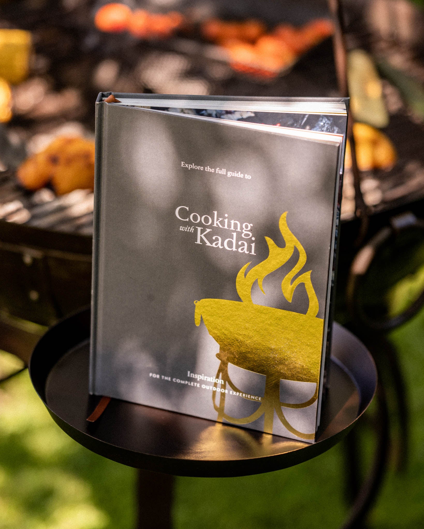 Cooking with Kadai live fire cookbook displayed on metal tray beside outdoor fire pit with glowing embers