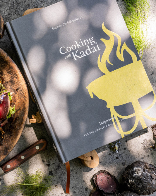 Cooking with Kadai live fire cookbook on outdoor surface beside grilled vegetables and knife