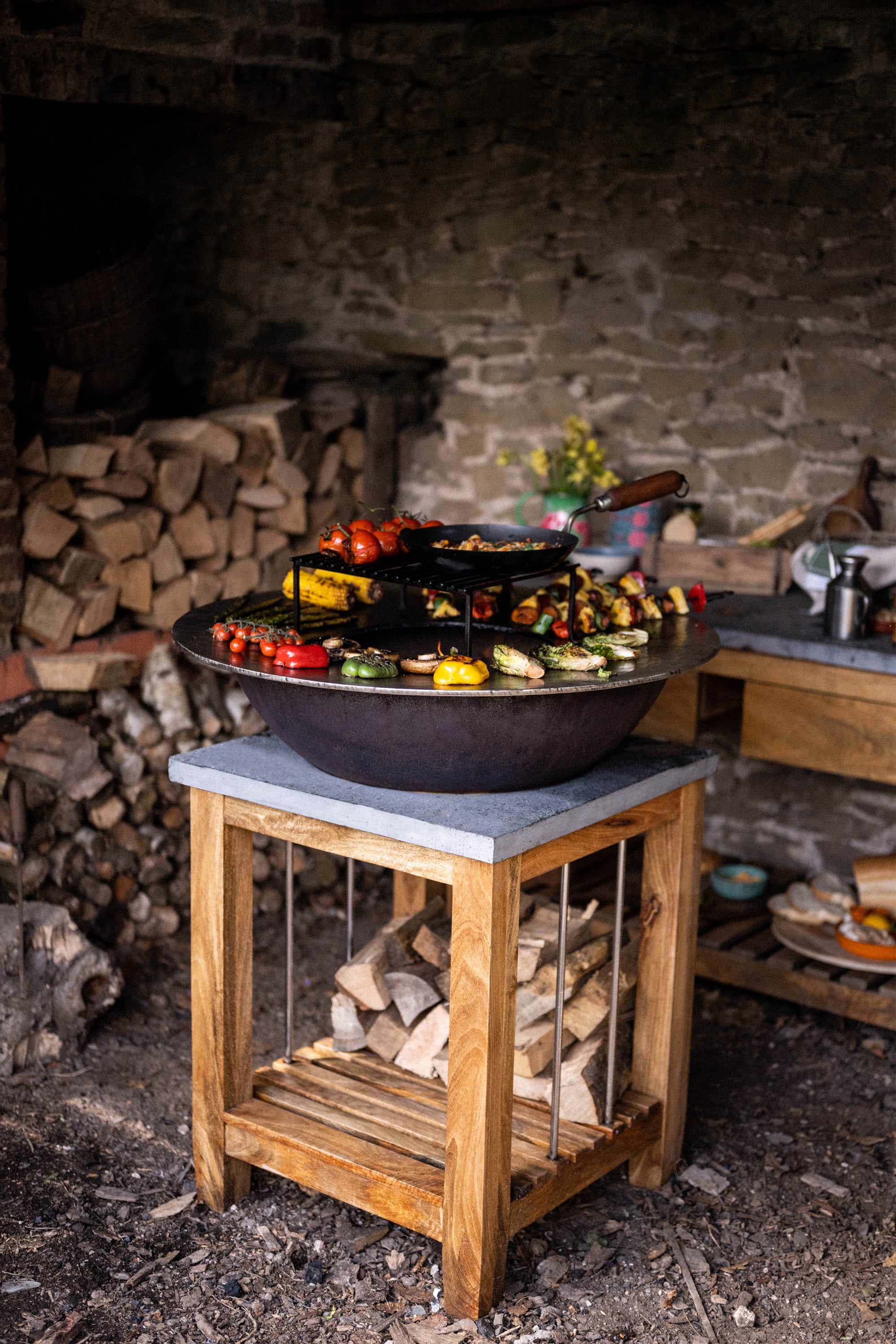 Kadai firepit with grilled vegetables and skewers on a wooden stand in a rustic outdoor kitchen setting