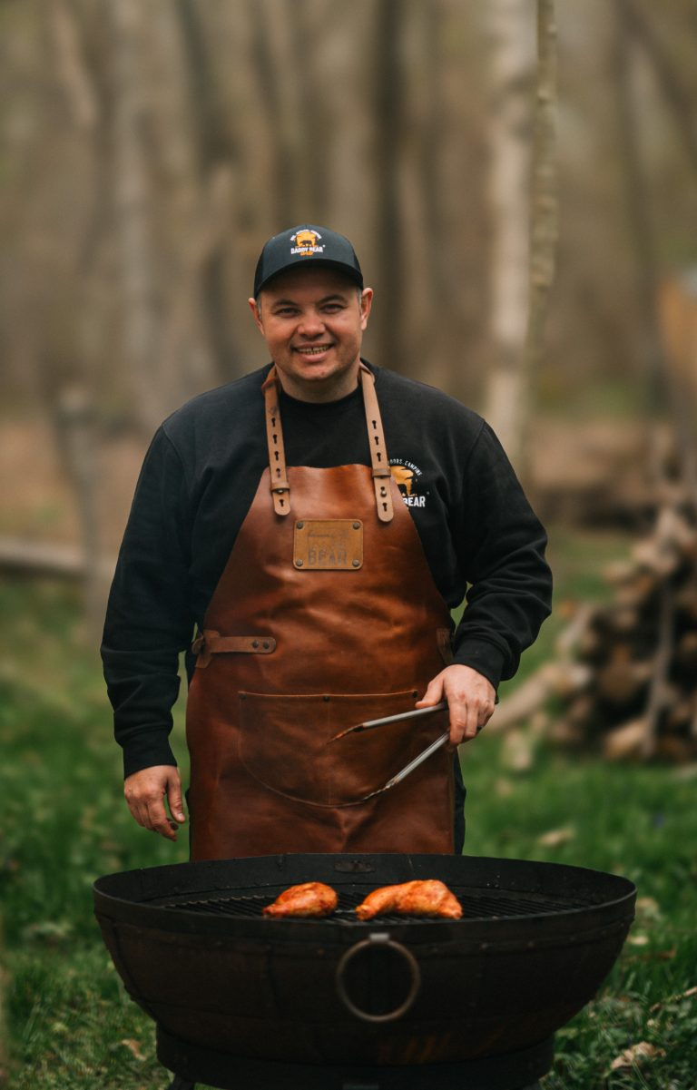 Man wearing a leather apron and cap, grilling outdoors on kadai firebowl with a blurred natural background